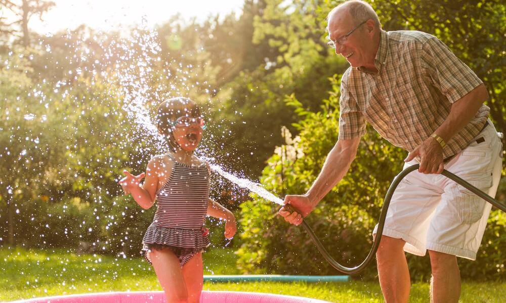 Young girl in striped swimsuit splashing in hose spray with grandfather